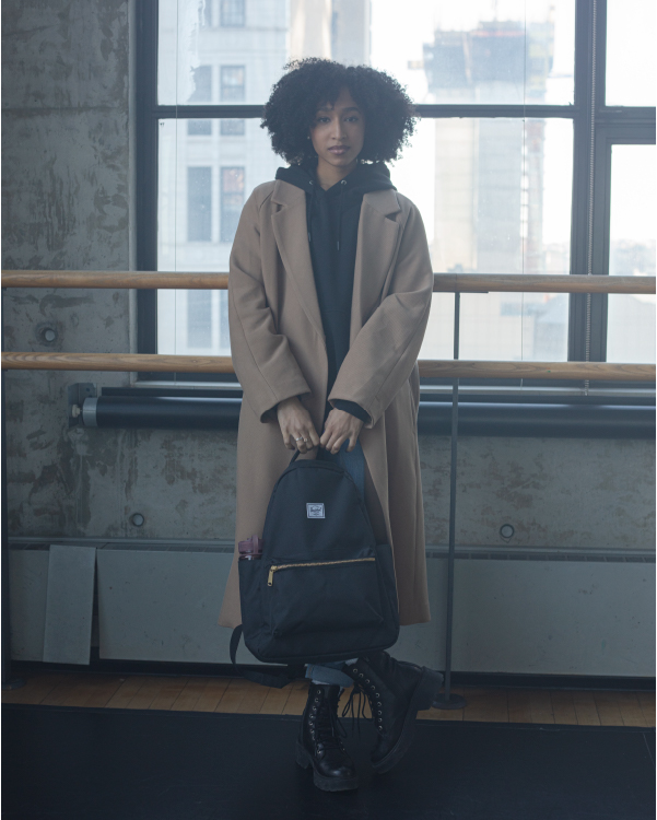 A woman is holding a Nova Backpack Mid-Volume in a ballet studio leaning against a waist bar